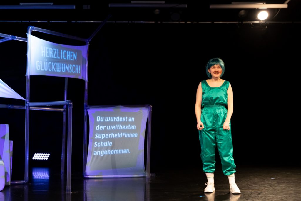 Camilla Pölzer,  a white, disabled cis woman, stands in the middle of the stage and smiles to the audience: she is happy to be accepted into the heroine school! Camilla wears a silky, shiny turquoise jumpsuit, a blue-green bob-cut wig and white tennis socks and sneakers. Next to her is a scaffolding with two stretched cloths. Text is projected onto the cloths: Congratulations! You have been accepted to the world's best superhero school.