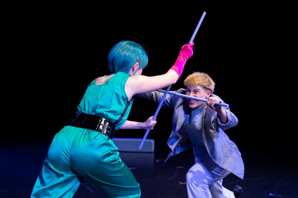 The two performers are in the middle of an intense stick fight. Camilla Przystawski is holding a stick with both hands in front of her body to defend herself. Her eyes are narrowed. She looks strained, but she is laughing. Camilla Pölzer is standing opposite her. She holds her stick vertically and presses it centrally against the other Camilla's, forcing her to her knees.
