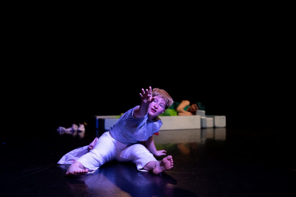Camilla Przystawski sits on the stage floor with her legs stretched out in front of her and reaches out a hand towards the audience as if grasping for something. Her gaze is expectant, her eyebrows raised and her mouth slightly open. The cool light casts shadows on her body. She wears short blonde hair, a shiny silver T-shirt and white trousers. In the blurred background, Camilla Pölzer lies curled up on foam blocks.