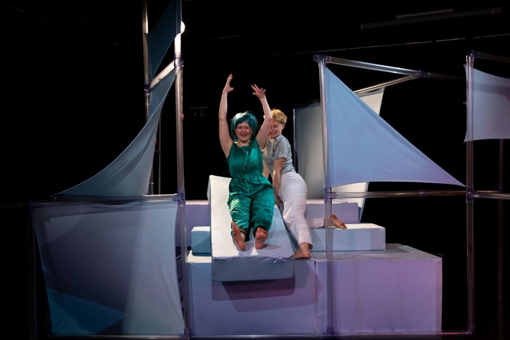 A scaffold with several levels of poles, stretched white cloths and white platforms. On it are the two performers. They have built a kind of slide with several foam blocks. Camilla Przystawski firmly holds the slide while Camilla Pölzer slides down with her arms stretched upwards. They both laugh playfully.