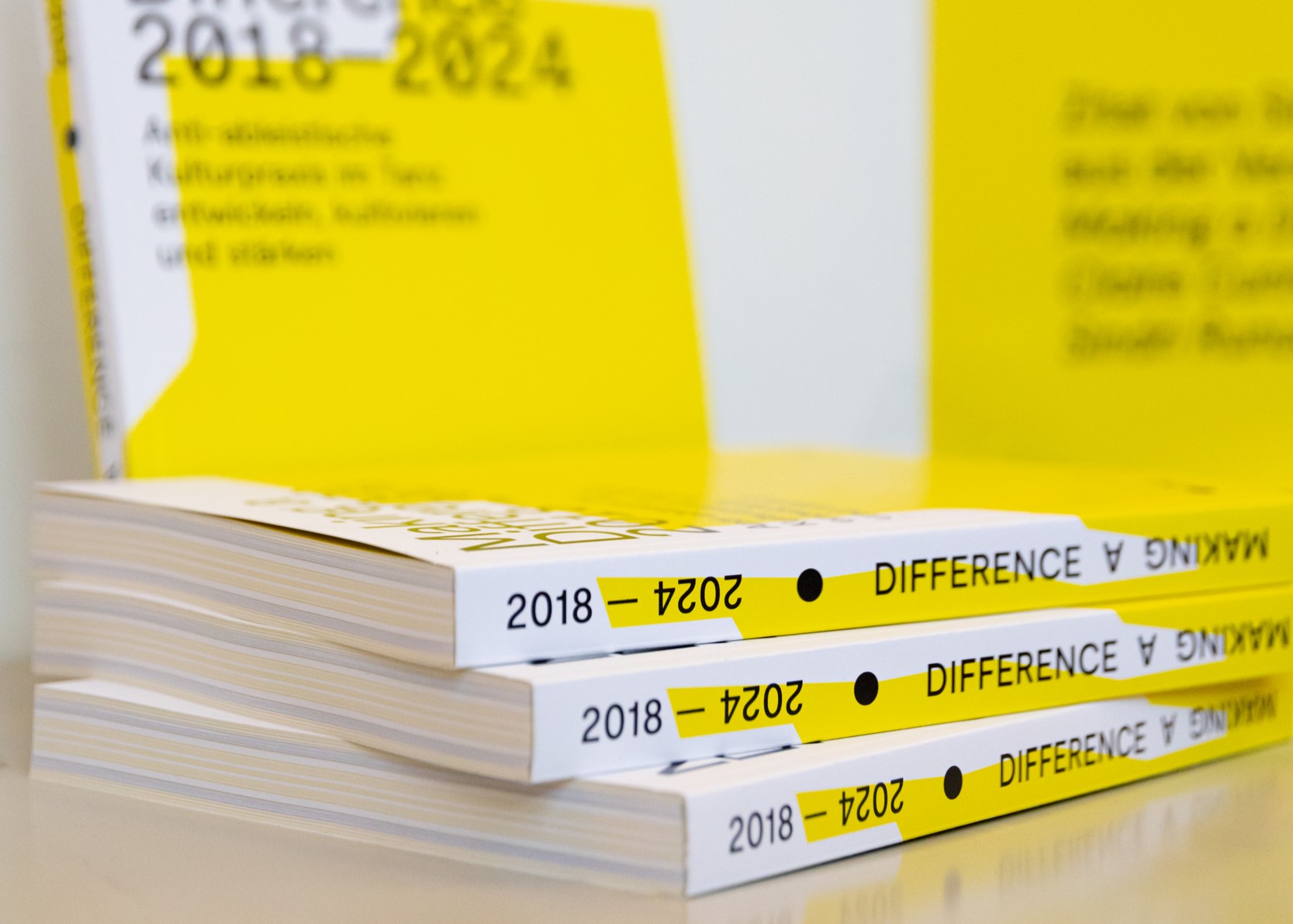Das Foto zeigt die Publikation als gedrucktes Buch. Im Vordergrund liegen drei Bücher aufeinandergestapelt, im Hintergund steht ein Buch aufrecht. Das Cover hat einen weißen und gelben Farbverlauf. Der Titel in schwarzer Schrift ist verschwommen.