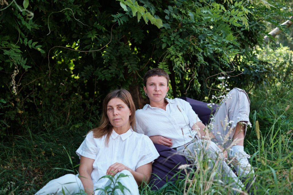 Juli, a female read, white person in her early 40s with dark blonde, long hair and Leo, a trans* non-binary, white person in their early 30s with short hair are sitting on or in front of a purple beanbag. They are sitting half in front of each other, half next to each other and leaning slightly towards each other. Leo is slightly leaning back and has one leg bent in front of them. They look straight and fixedly into the camera. Juli is leaning against the beanbag with her left hand resting on her bent knee and her head turned slightly to the side. She is also looking directly into the camera. Both are wearing (by chance) white, short-sleeved shirts and light-coloured trousers. Plants are growing around them. In front, tall grass partially covers their legs, while the background is fully covered by a green climbing plant blocking out the daylight.