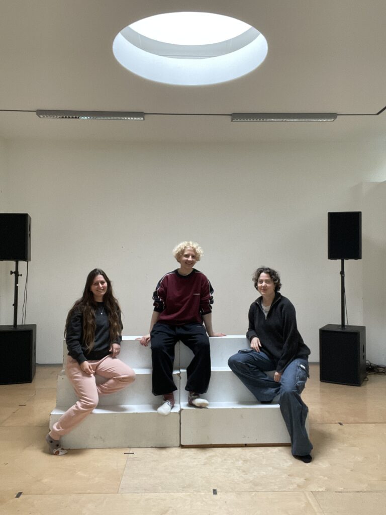 The photo was taken in a rehearsal studio. In the middle is a three-step white model staircase. Ariana, Silva and Lee sit on a three-step white model staircase in the middle. On the left sits Ariana, slightly turned towards the other two, smiling into the camera. She is German-Iranian and in her late 20s. She wears comfortable sportswear and her long brown hair down. Next to her, in the middle of the top step, sits Silva (30). Silva is a white non-binary person with short blond curls in comfortable sportswear. They prop themselves up with both hands and laugh into the camera. To Silva's right sits Lee. Like Ariana, Lee is a little turned towards the other two, one elbow casually resting on the top step. Lee is 30, a non-binary/trans person and has short curly brown hair that is already graying a bit at the roots. Lee also wears comfortable sports clothes and smiles into the camera. In the ceiling above them is a round window through which light falls on the three of them. In the background there are two black loudspeakers on the left and right.