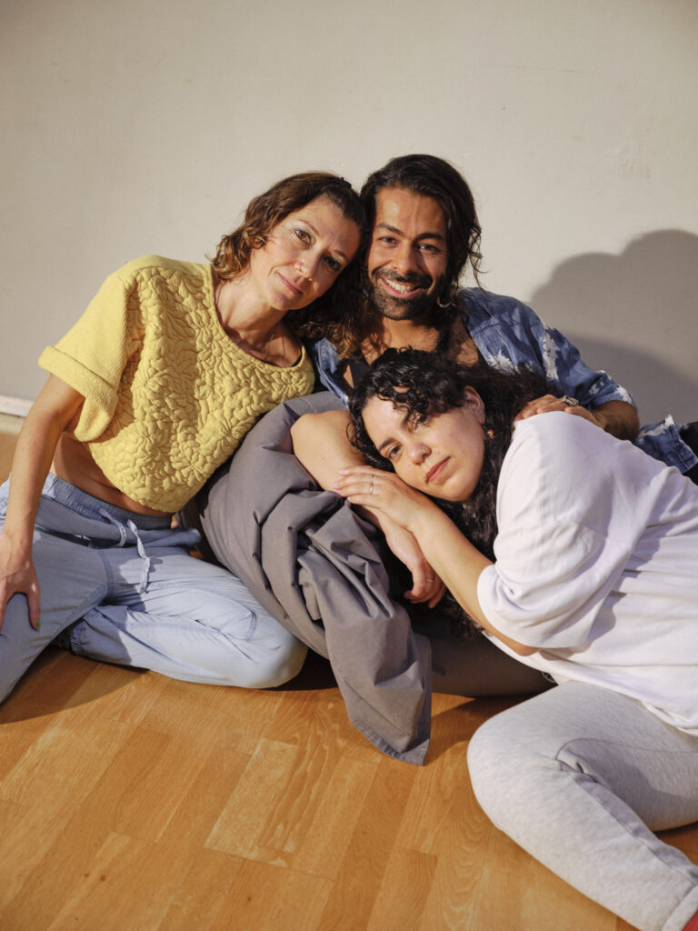 Three adults sit close together on a wooden floor in front of a light-colored wall. One person rests their head on the shoulder of another, while a third person lies in front of them, resting their head on their arm. 